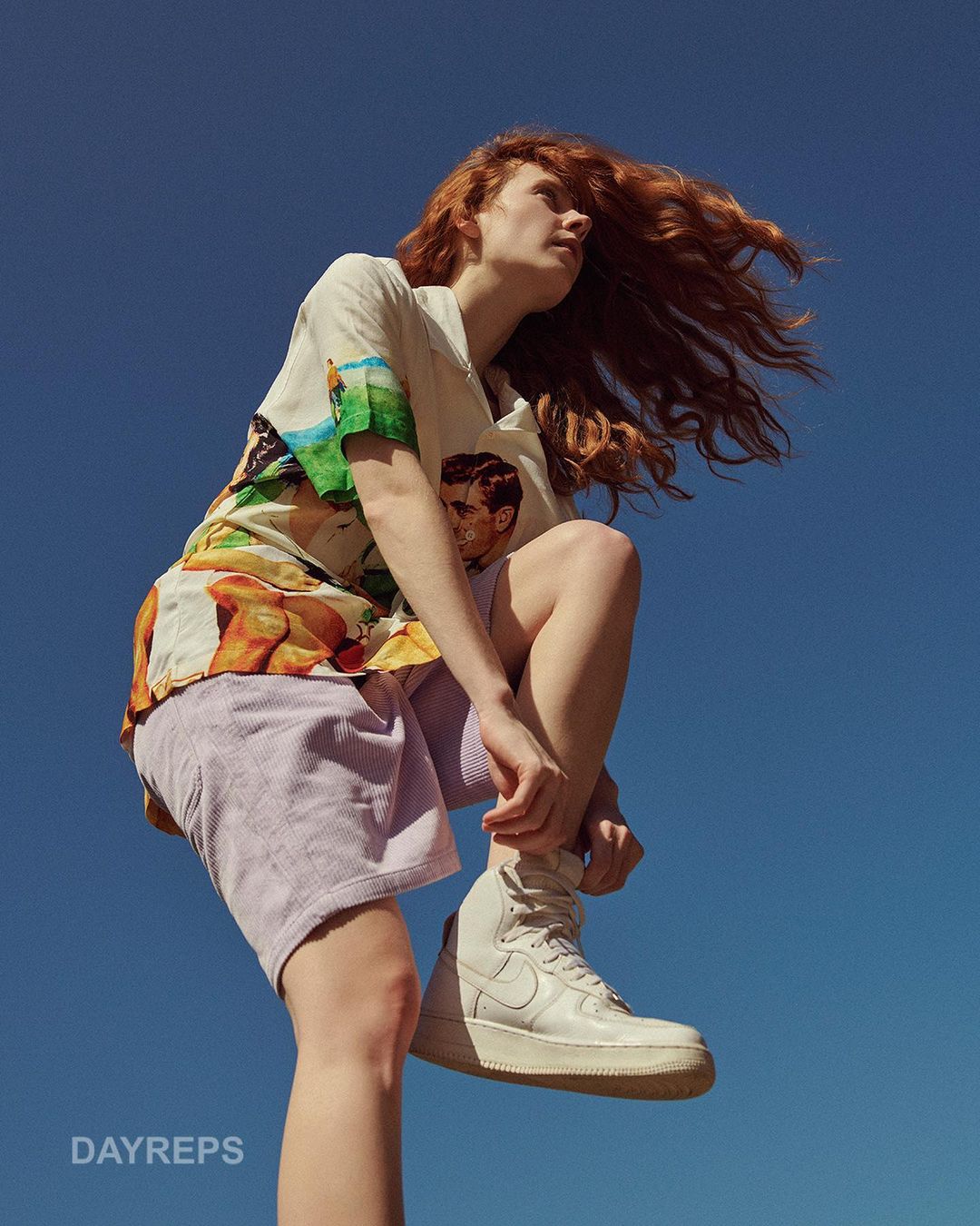 Young woman with red hair wearing a patterned shirt and shorts tying a white sneaker against a vivid blue sky backdrop.