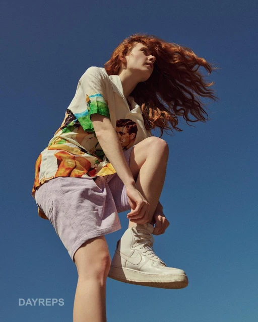 Young woman with red hair wearing a patterned shirt and shorts tying a white sneaker against a vivid blue sky backdrop.