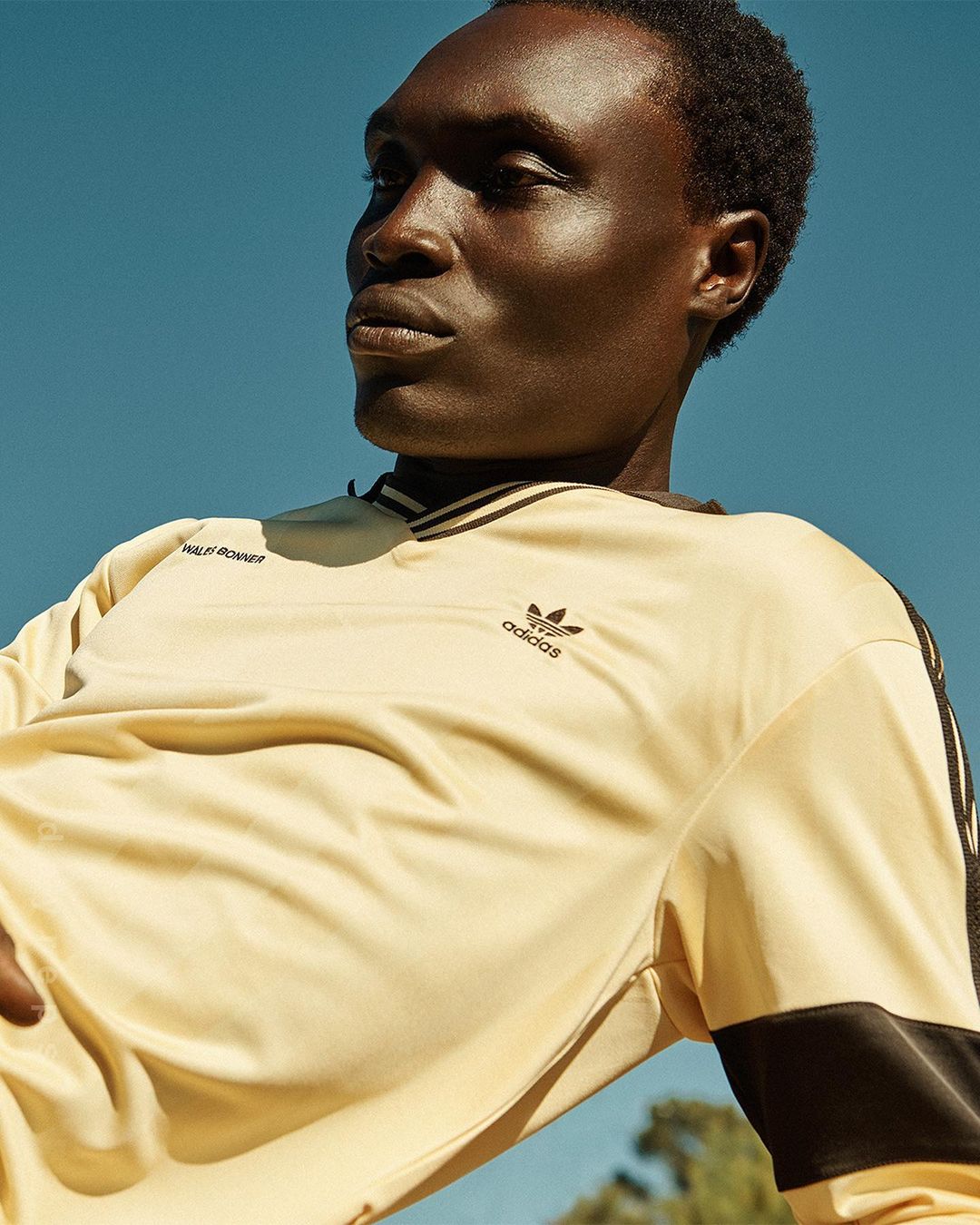 A dark-skinned male model poses outdoors in a cream-colored Adidas long-sleeve shirt against a bright blue sky backdrop.