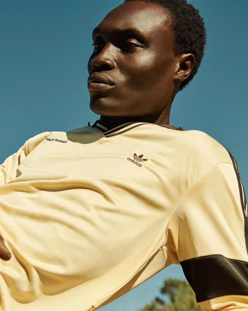 A dark-skinned male model poses outdoors in a cream-colored Adidas long-sleeve shirt against a bright blue sky backdrop.
