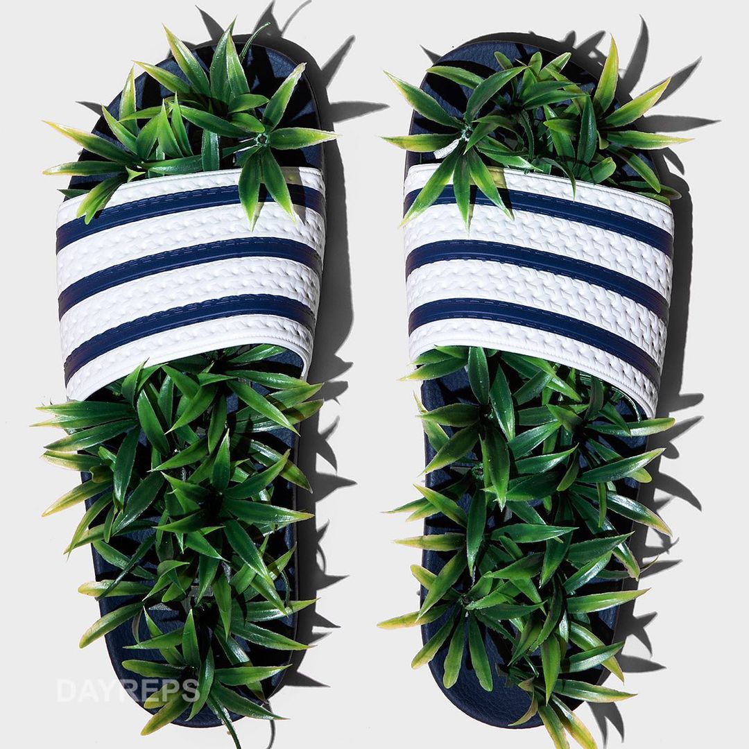 Pair of navy and white striped sandals with green leafy plants sprouting from them, set on a neutral-toned surface, studio shot.