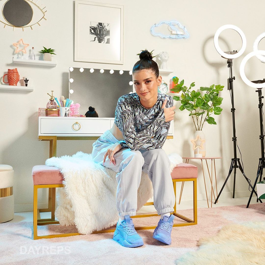 Young woman sits on a pink and gold bench in front of a vanity with ring lights and wall decorations in a staged interior.