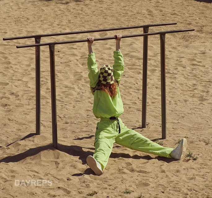 A woman hangs from parallel bars in a playground, wearing a green tracksuit and checkerboard hat, her legs extended. Bright, sunny day.