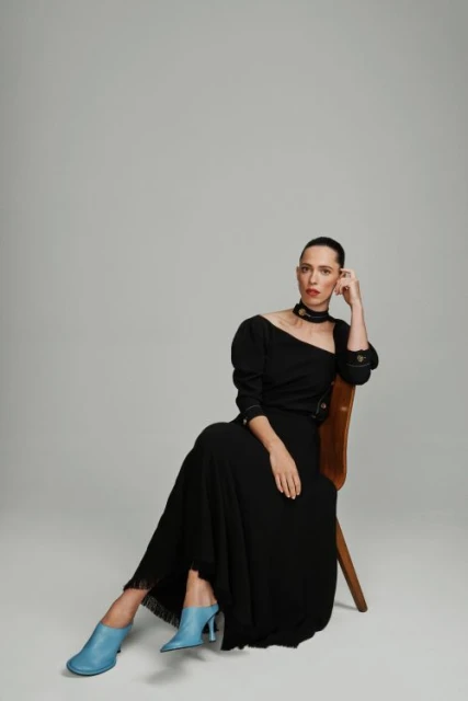 Elegant woman in a black dress sits gracefully on a wooden chair against a neutral background in a fashion photography setting.