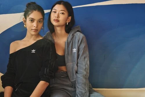 Two fashionable adult women model Adidas apparel against a blue backdrop in a studio. One wears a black top, the other a grey tracksuit.