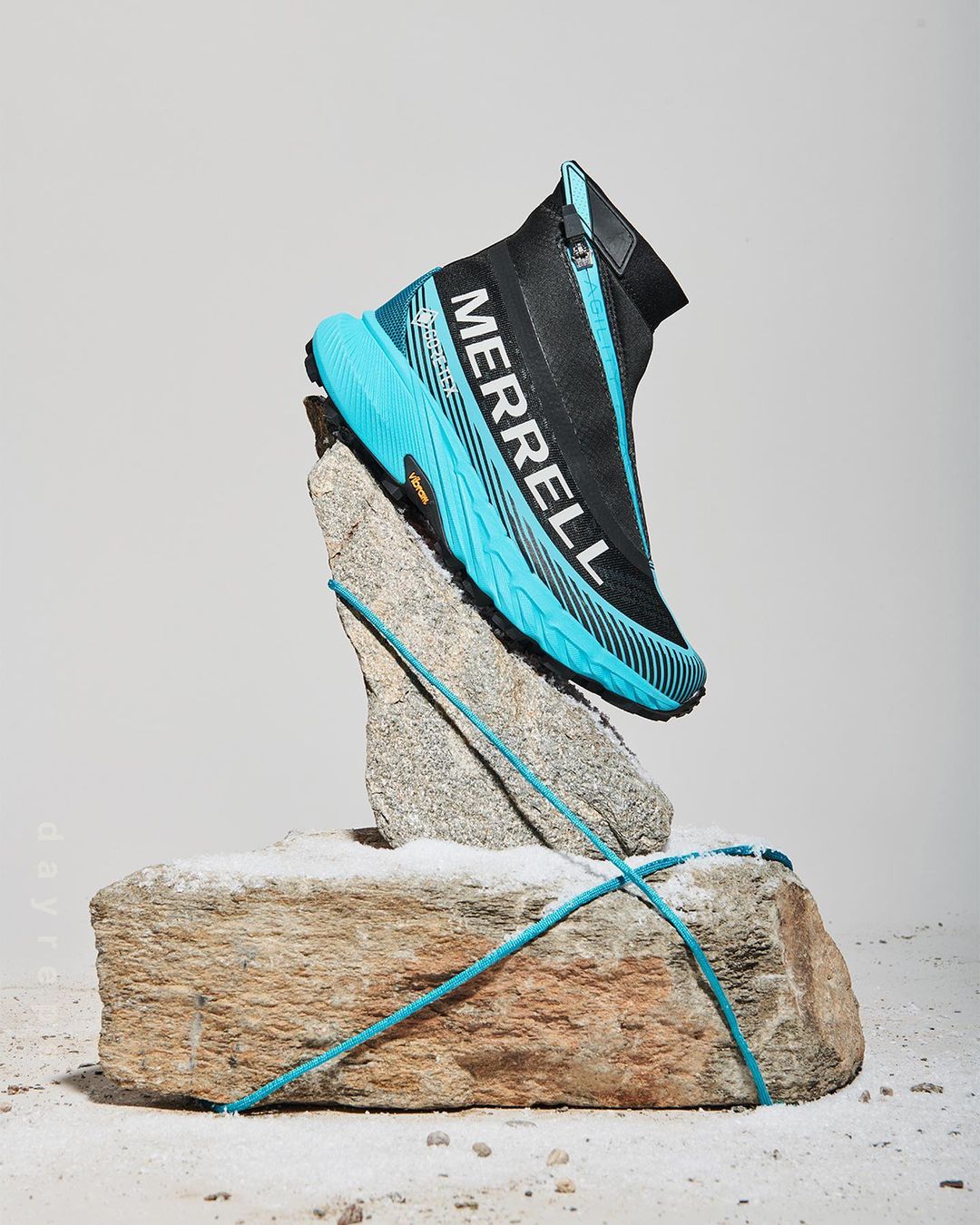 A turquoise and black Merrell zip-up sneaker is displayed on a rough grey rock in a studio setting with a bright white background.