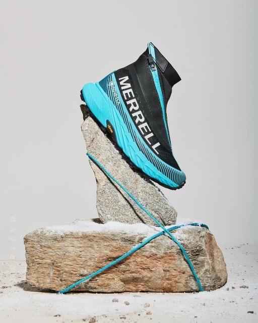 A turquoise and black Merrell zip-up sneaker is displayed on a rough grey rock in a studio setting with a bright white background.