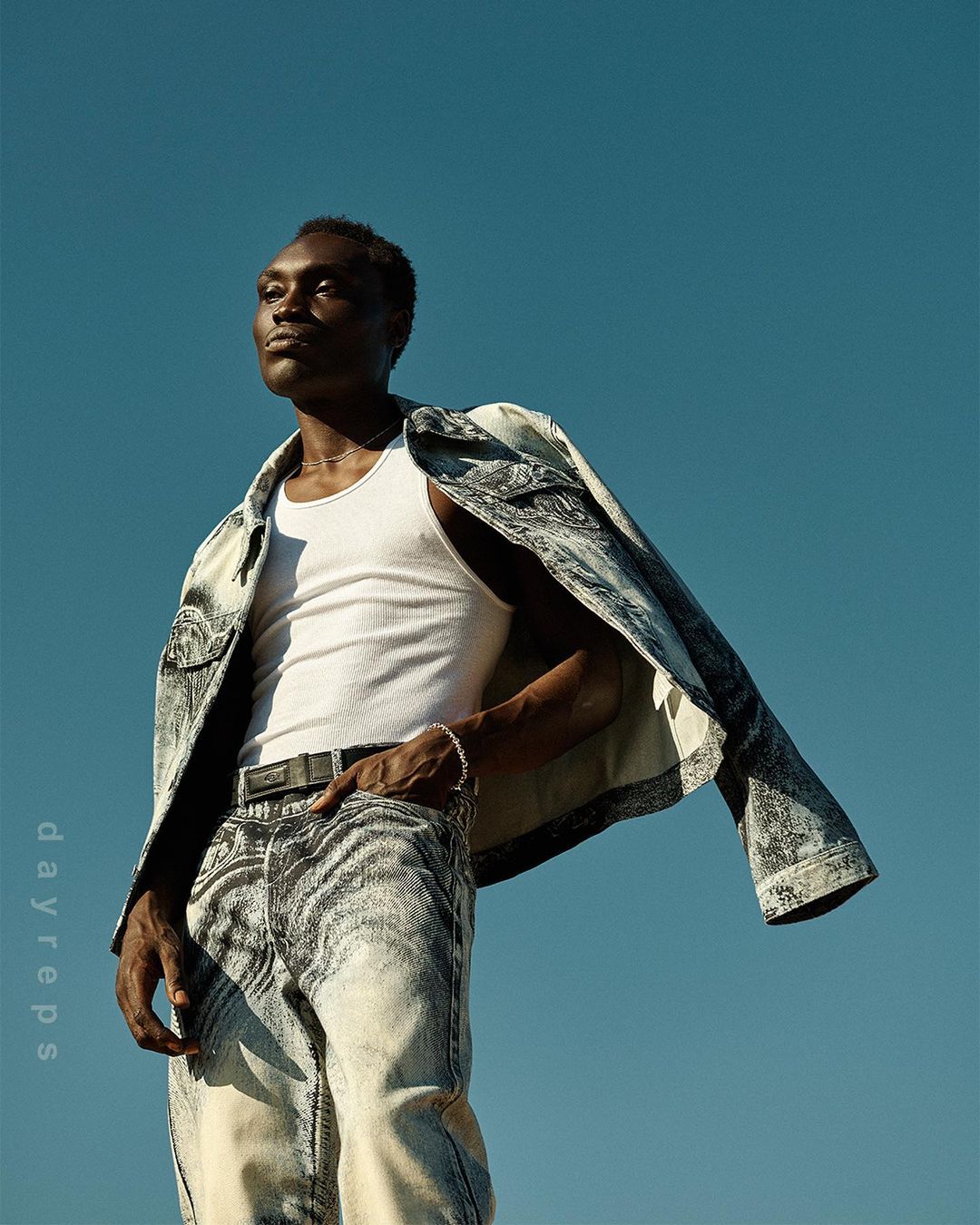 Dark-skinned man in acid-wash denim jacket and jeans poses against a clear blue sky, striking a confident pose with a neutral expression.