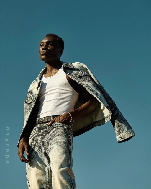 Dark-skinned man in acid-wash denim jacket and jeans poses against a clear blue sky, striking a confident pose with a neutral expression.