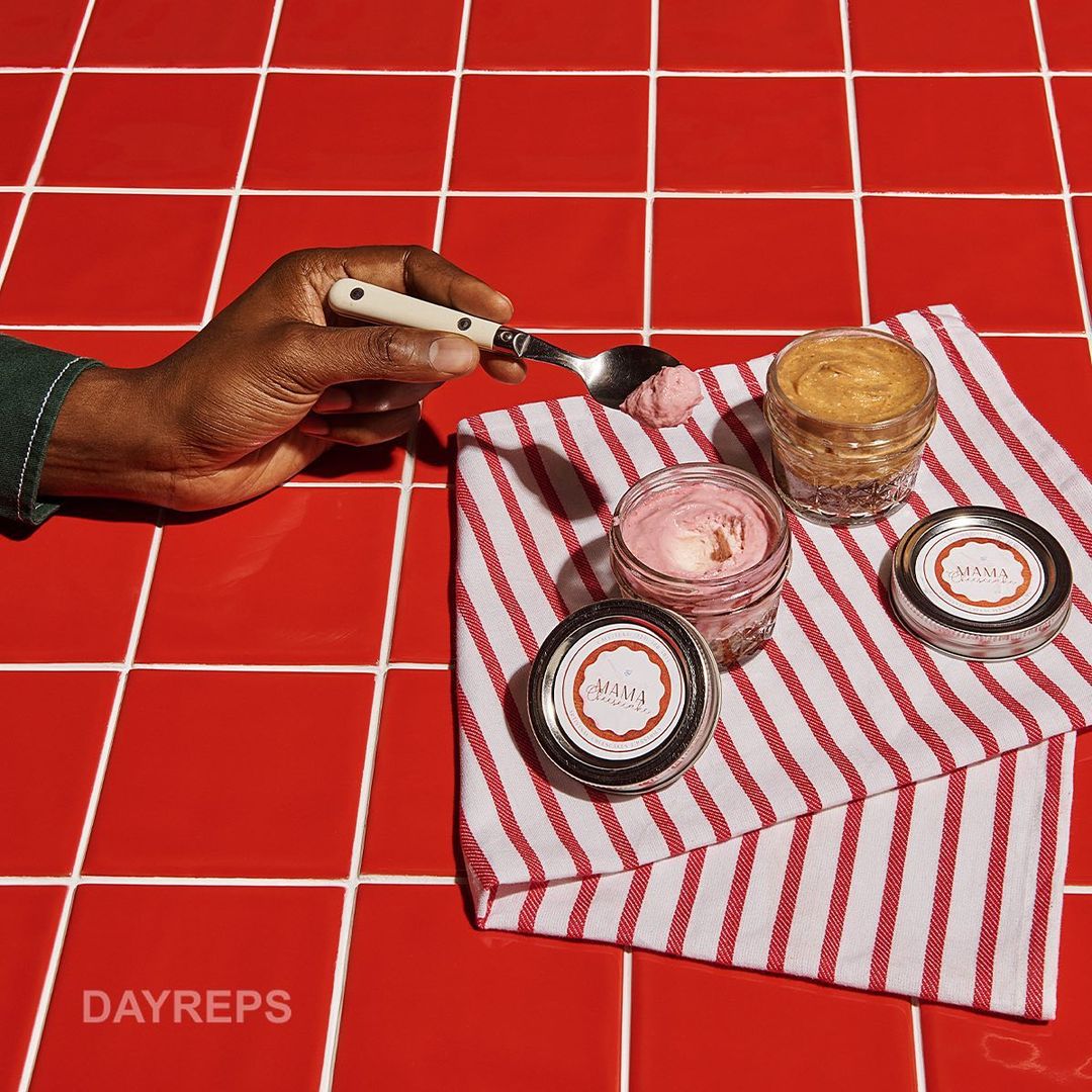 Close-up of a hand holding a spoon with pink ice cream next to glass jars with lids and a red-and-white striped towel on red tiles.