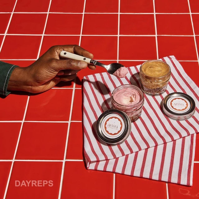 Close-up of a hand holding a spoon with pink ice cream next to glass jars with lids and a red-and-white striped towel on red tiles.