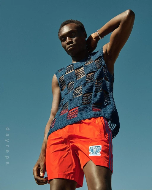 Young Black male model poses against a blue sky wearing a blue woven top and bright orange Adidas shorts in an outdoor fashion shot.