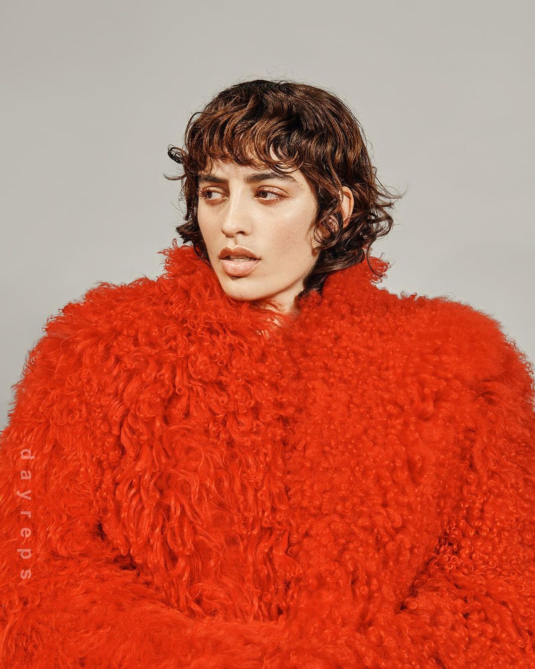 Portrait of a woman with short wavy brown hair wearing a bright red, textured faux fur coat against a solid grey backdrop in a studio.