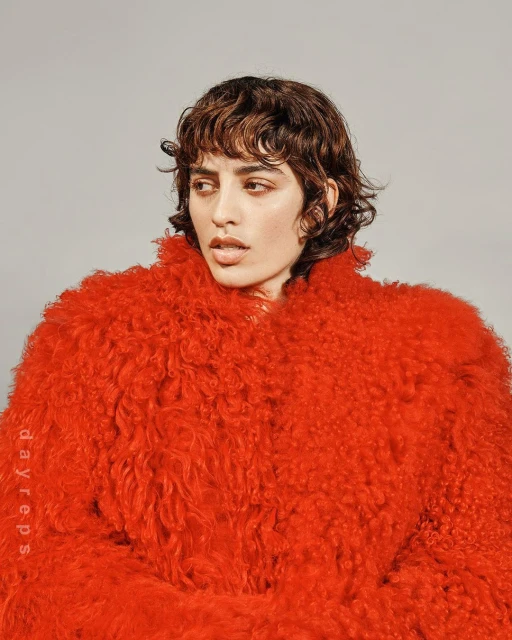 Portrait of a woman with short wavy brown hair wearing a bright red, textured faux fur coat against a solid grey backdrop in a studio.