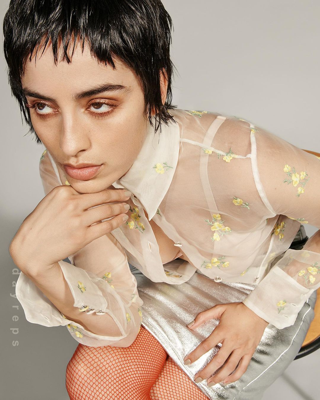 Portrait of a young woman with short hair wearing a sheer floral blouse and silver skirt, sitting against a neutral background.