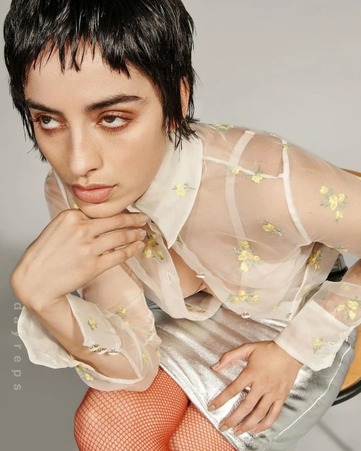 Portrait of a young woman with short hair wearing a sheer floral blouse and silver skirt, sitting against a neutral background.
