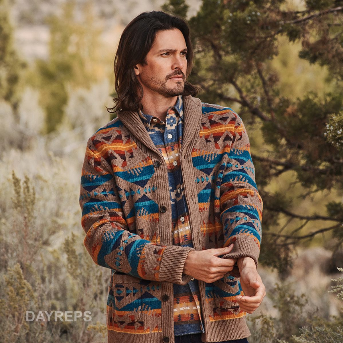 Man wearing a patterned cardigan and collared shirt stands in a natural outdoor setting with neutral colours.  
