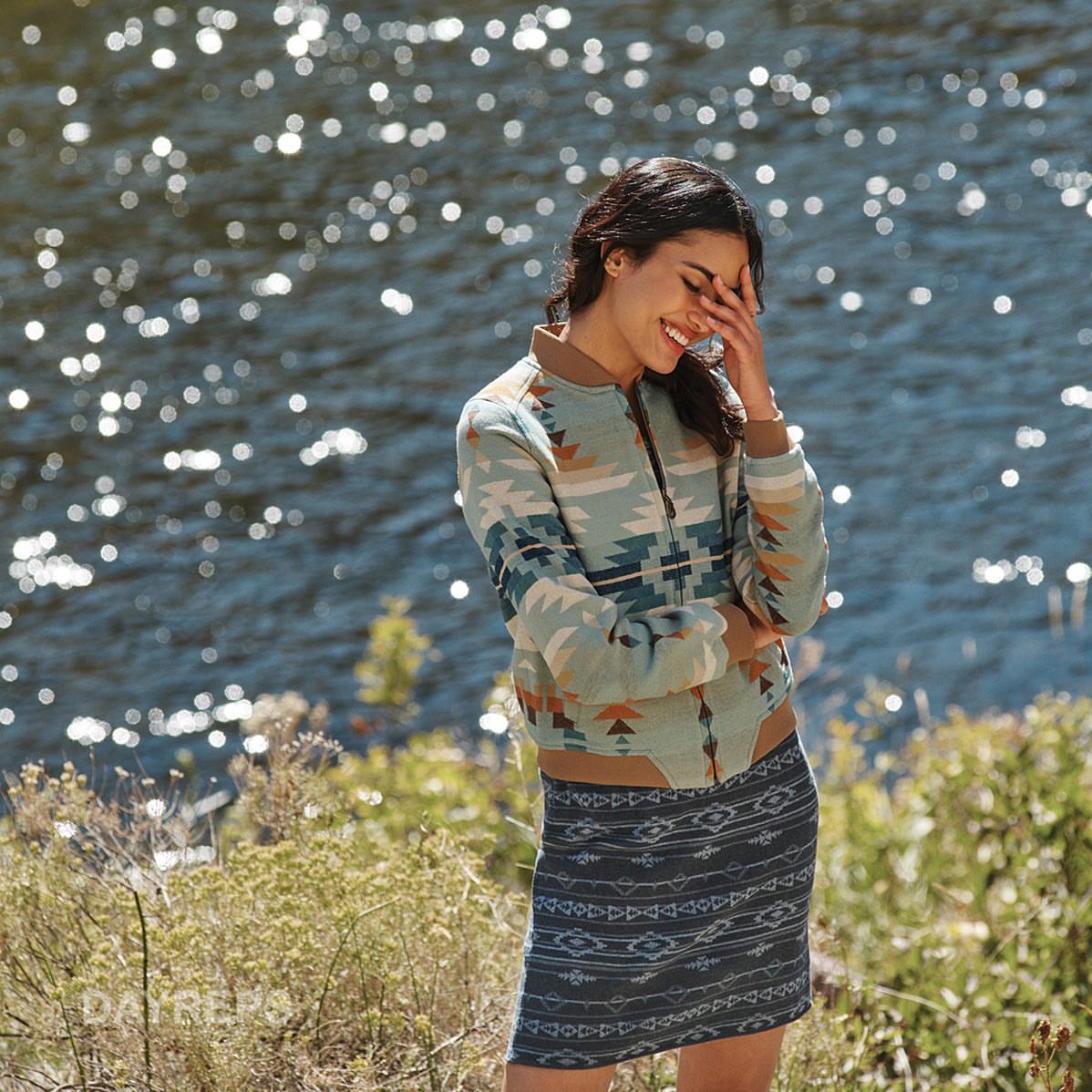 A woman in a patterned jacket and skirt smiles with hand near her face, with a blurred river and greenery in the background.