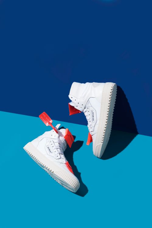 Two white high-top sneakers from OFF-WHITE angled on a split-tone blue background, showing their unique designs and orange tags.