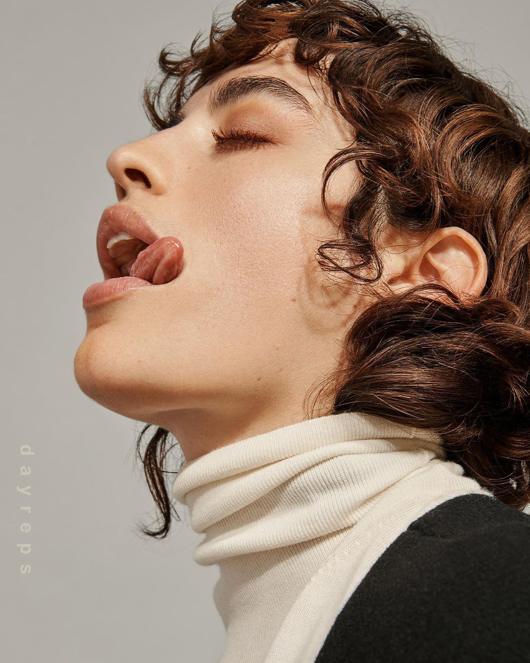 Profile portrait of a woman with short brown curly hair, wearing a cream turtleneck, eyes closed, mouth open, and tongue extended.