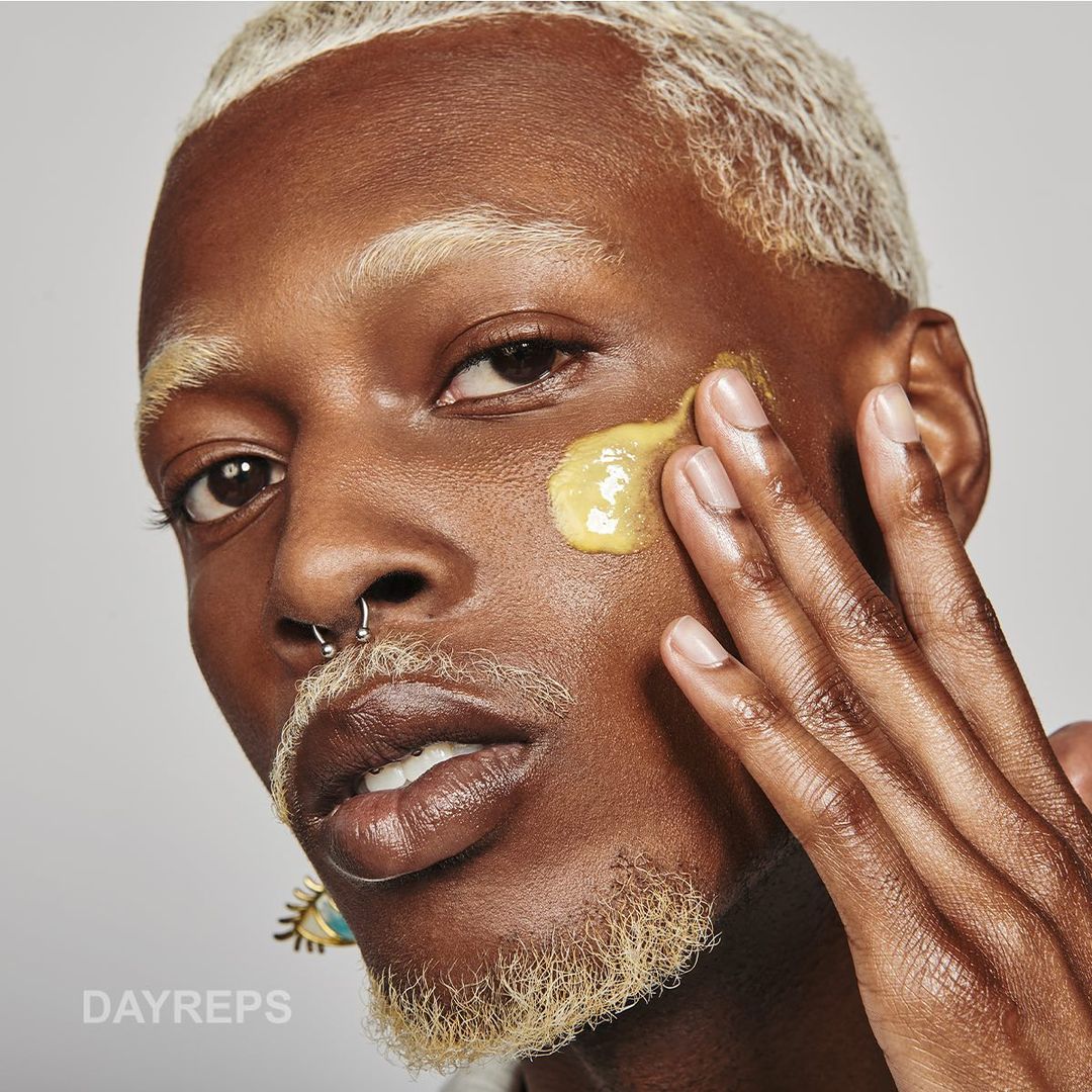 Close-up portrait of a person with bleached blonde hair and eyebrows applying a yellow cream to their cheek by Molly Cranna