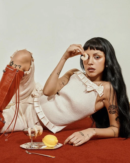 Woman in cream outfit poses with shrimp and a martini on a red surface in a studio setting; she is looking at the camera.