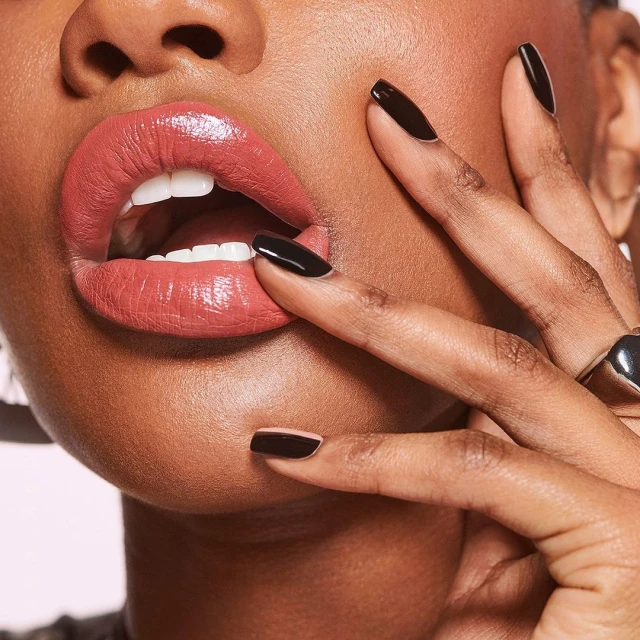 Close-up of an adult woman with an open mouth and a salmon-colored lipstick touching her lips with fingers wearing black nail polish.