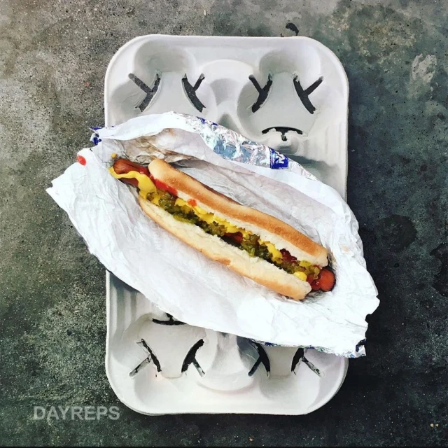 Overhead shot of a hot dog with mustard and relish in a foil wrapper on a white cardboard drink carrier sitting on a gray surface.