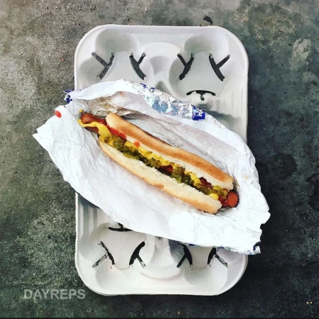 Overhead shot of a hot dog with mustard and relish in a foil wrapper on a white cardboard drink carrier sitting on a gray surface.