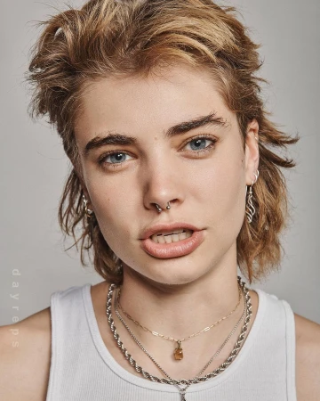 Close-up portrait of a person with blonde shaggy hair, septum piercing, and layered silver necklaces wearing a white tank top