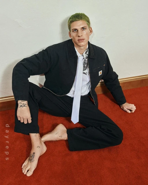 A trendy adult male with green hair and tattoos poses barefoot on a red carpet wearing black jacket and pants, and a white tie against a neutral wall.