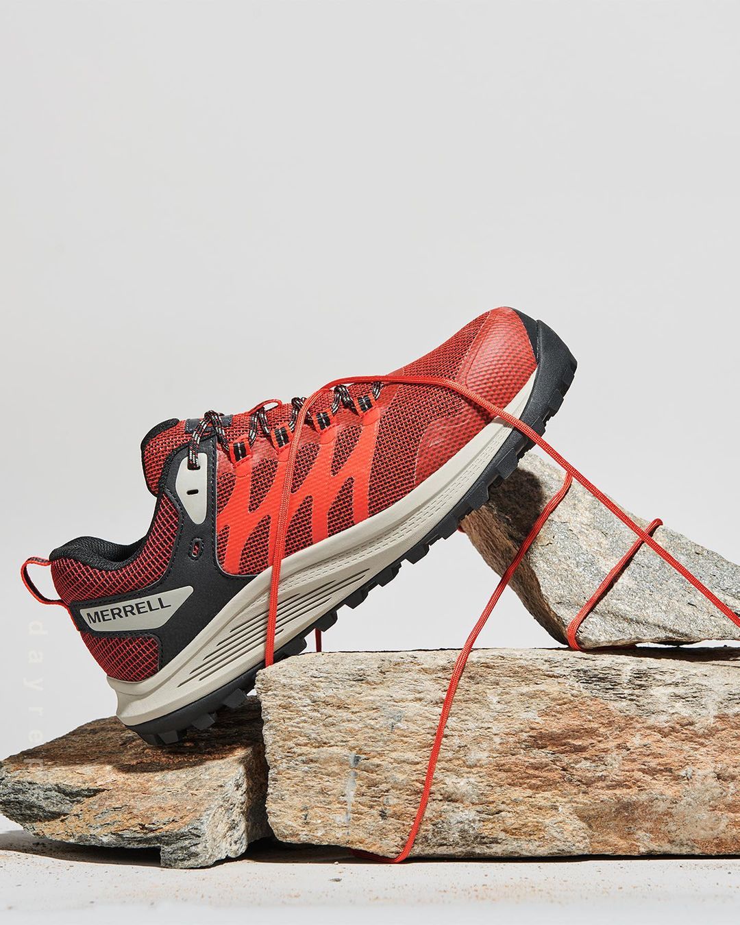 Red and black Merrell trail running shoe, studio shot, resting on textured grey rocks against a white backdrop.