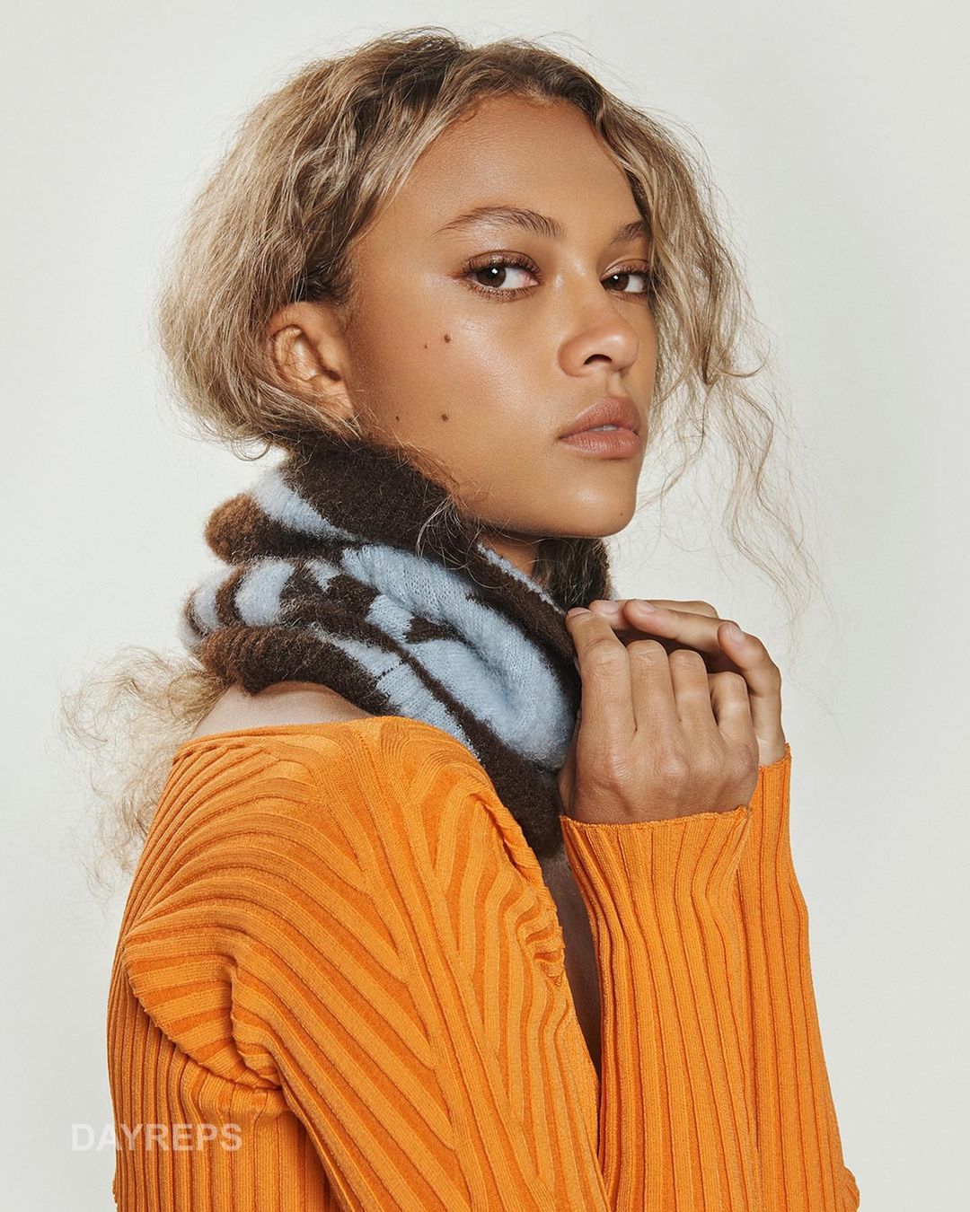Close-up of a woman with curly blonde hair, wearing a blue and brown scarf and orange shirt, looking calmly at the camera in a photo studio.