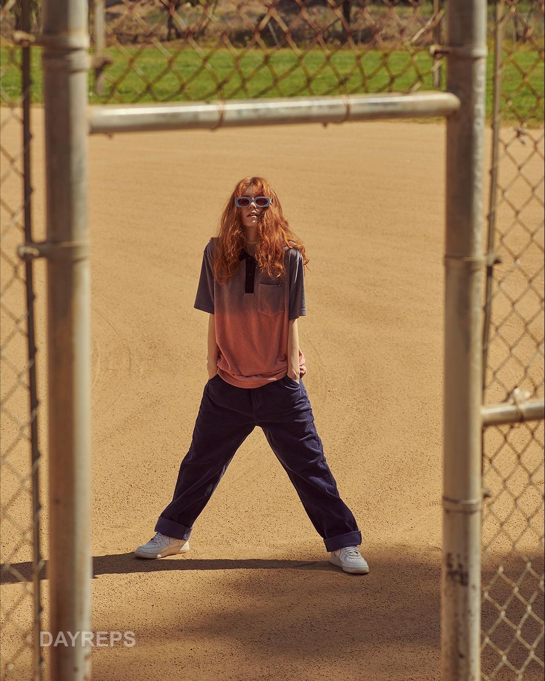 A red-haired woman in a polo shirt and blue pants stands in a baseball field, framed by a chain-link fence in portrait orientation.