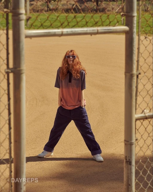 A red-haired woman in a polo shirt and blue pants stands in a baseball field, framed by a chain-link fence in portrait orientation.
