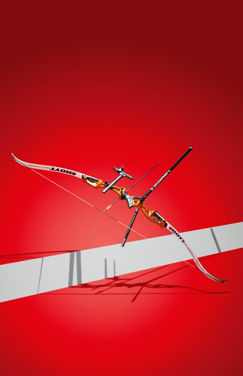A modern recurve bow in a studio setting, angled with metallic and white accents, set against a vibrant red background with a white stripe.