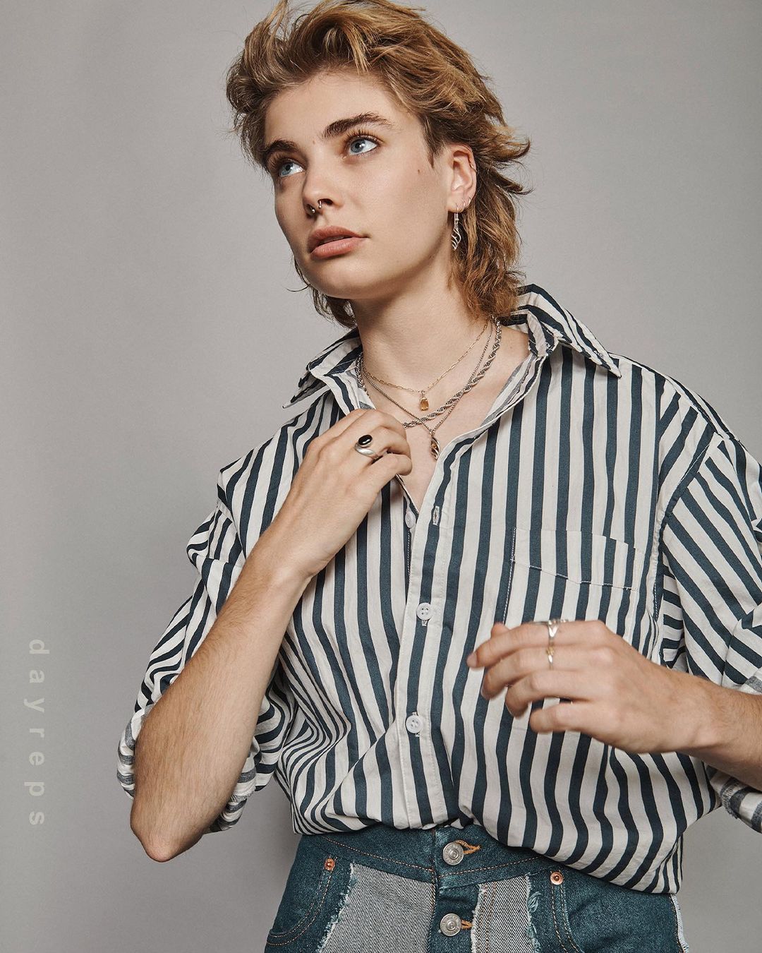 Portrait of an adult with light skin, wearing a striped shirt and multiple necklaces, looking off-camera against a neutral background.