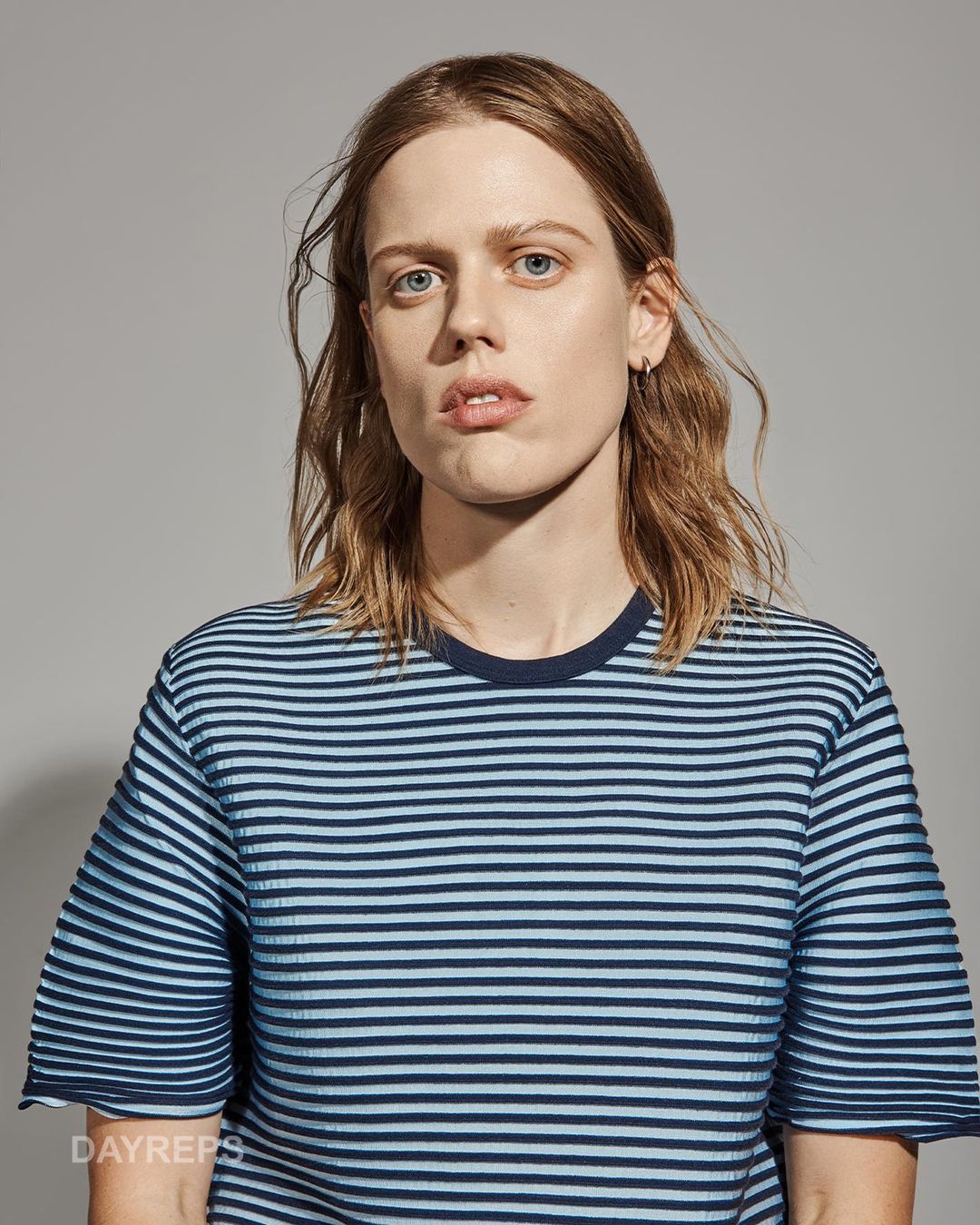 Model with wavy brown hair wearing a blue striped t-shirt against a plain grey background