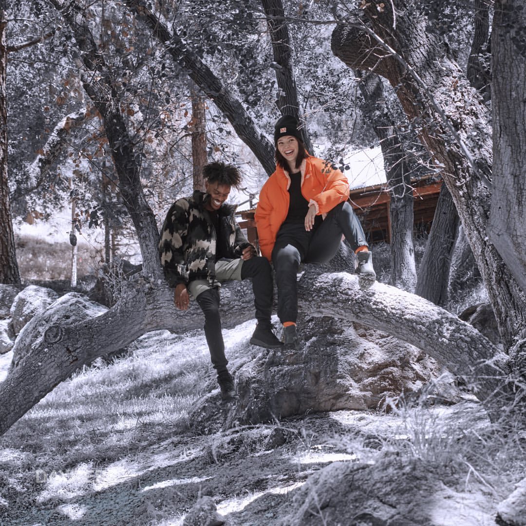 Two people are sitting on a tree trunk in the forest. The man is wearing a camouflage jacket and the woman is wearing an orange jacket.