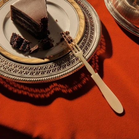Slice of chocolate cake on decorative plate with fork, presented on an orange fabric tablecloth with natural light.