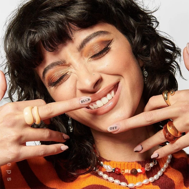 Smiling woman with dark hair, brown eye makeup, wearing beaded jewelry and rings, holding hands up with tattoo 'BUCK OFF'.