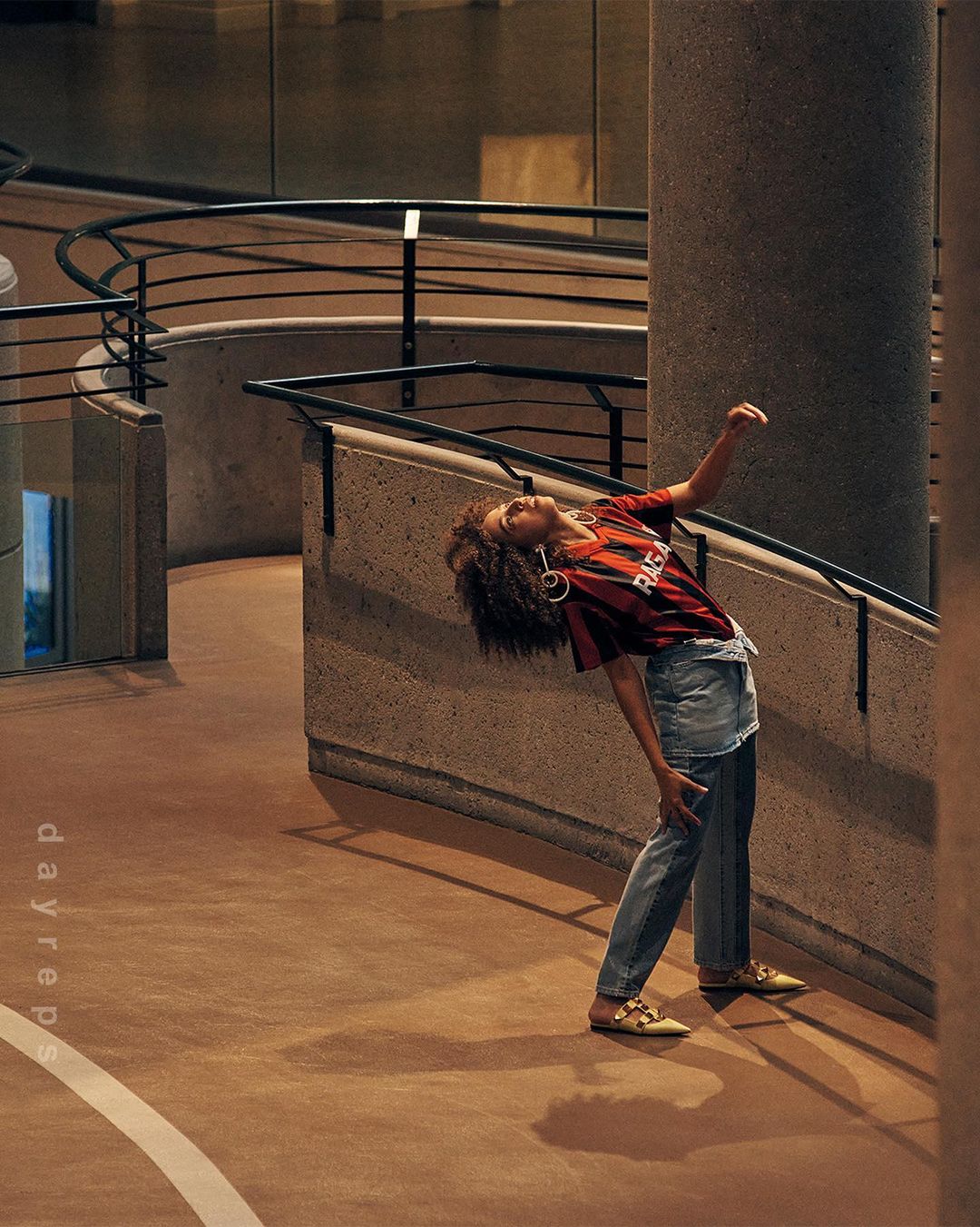 Woman poses in jeans and a graphic tee inside a modern building featuring concrete walls, a staircase, and black metal railings.