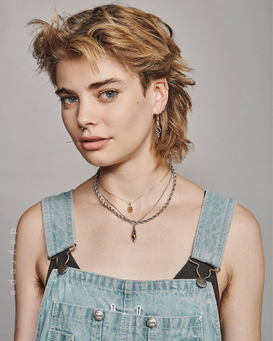 Close-up portrait of a young adult female with a mullet hairstyle wearing denim overalls against a grey backdrop.