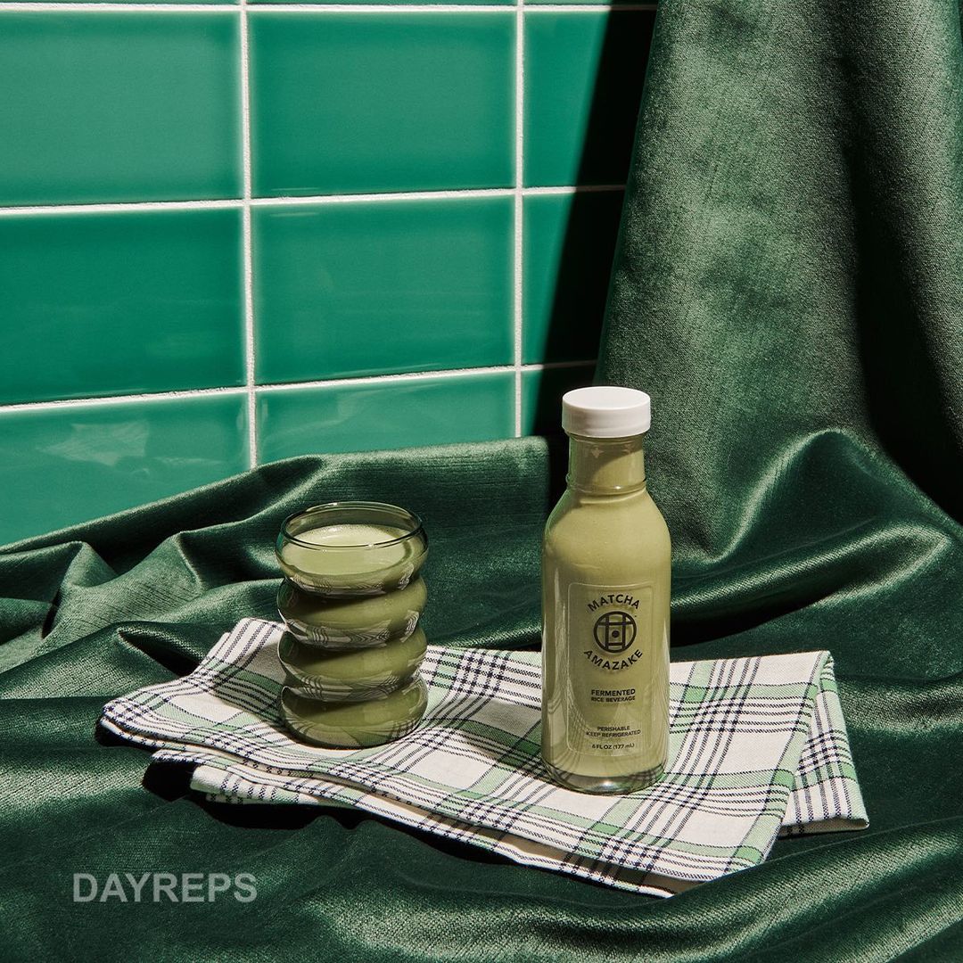Matcha Amazake bottle next to glass on checkered cloth against a background of green tiles and draped green velvet fabric.