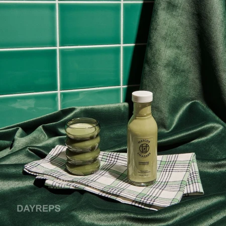 Matcha Amazake bottle next to glass on checkered cloth against a background of green tiles and draped green velvet fabric.
