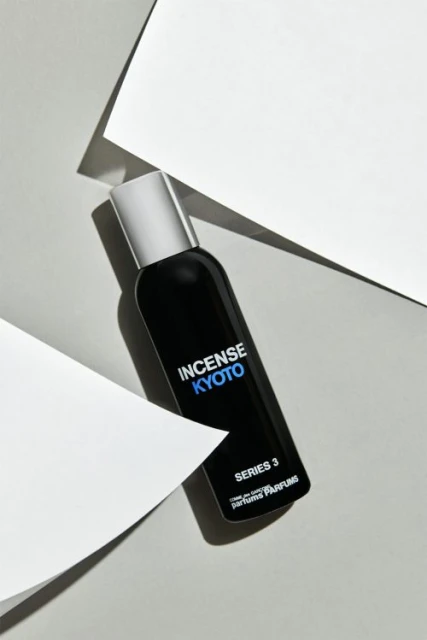 A black perfume bottle with a silver cap stands out against white geometric shapes and a soft grey background in a beauty still life.