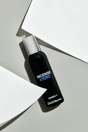 A black perfume bottle with a silver cap stands out against white geometric shapes and a soft grey background in a beauty still life.