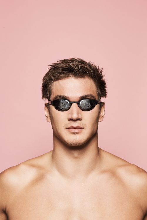 Close-up studio shot of a male swimmer wearing dark goggles against a pink background. Focus on the swimmer and equipment.