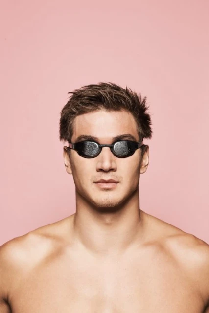 Close-up studio shot of a male swimmer wearing dark goggles against a pink background. Focus on the swimmer and equipment.