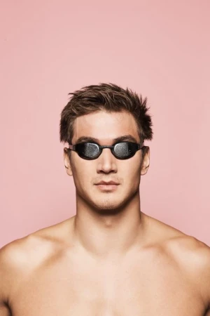 Close-up studio shot of a male swimmer wearing dark goggles against a pink background. Focus on the swimmer and equipment.